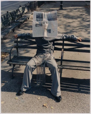 Person wearing a newspaper mask sitting on a park bench in sunlight. Figure is wearing a grey jacket and pants with black shoes.