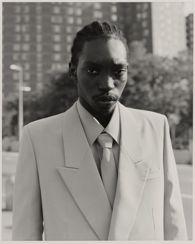 Close-up portrait of a Black man in a suit and tie, standing outdoors in an urban setting, with residential buildings in the background.