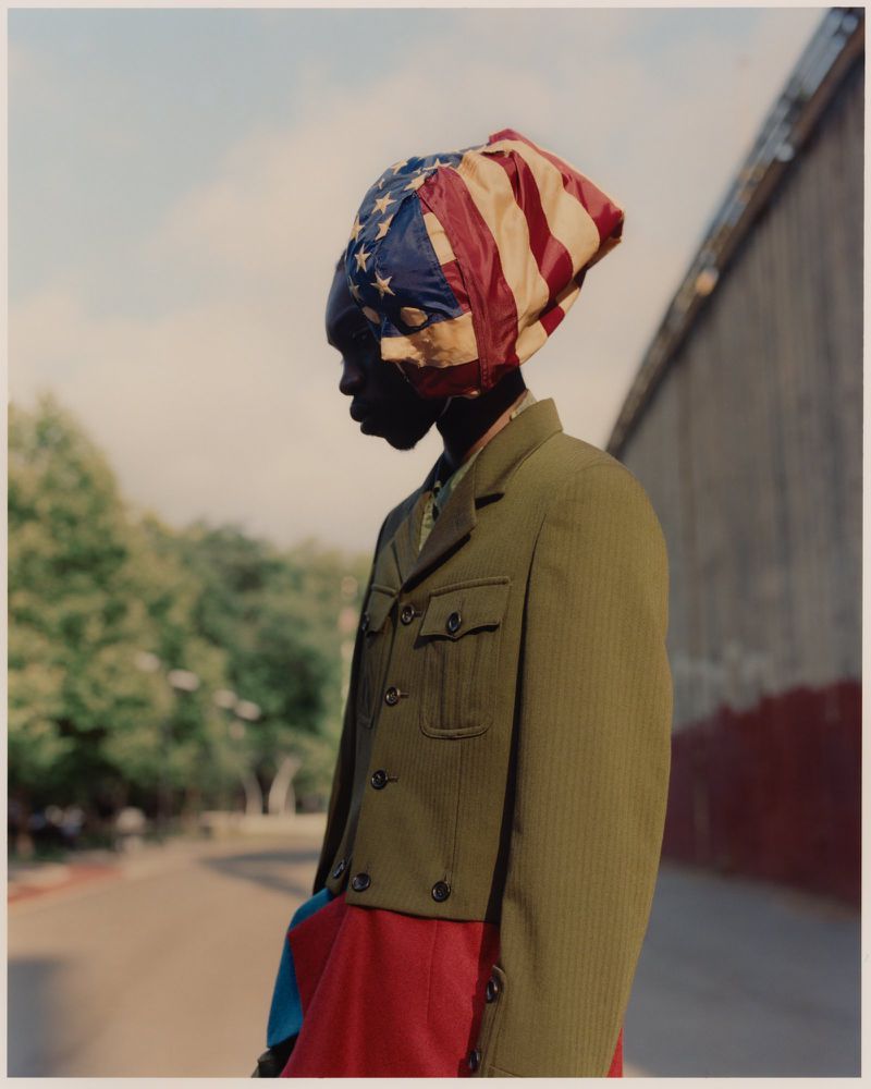 Adult stands on a street under blue sky wearing an olive drab jacket and a colorful head covering with American flag motif.