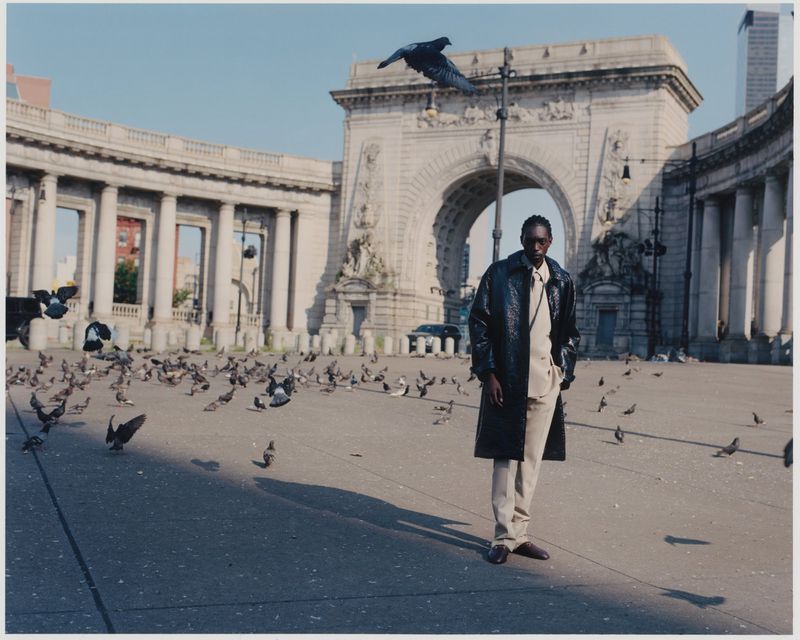 An adult male fashion model poses in front of the Manhattan Bridge Archway, New York, with numerous pigeons in the foreground.