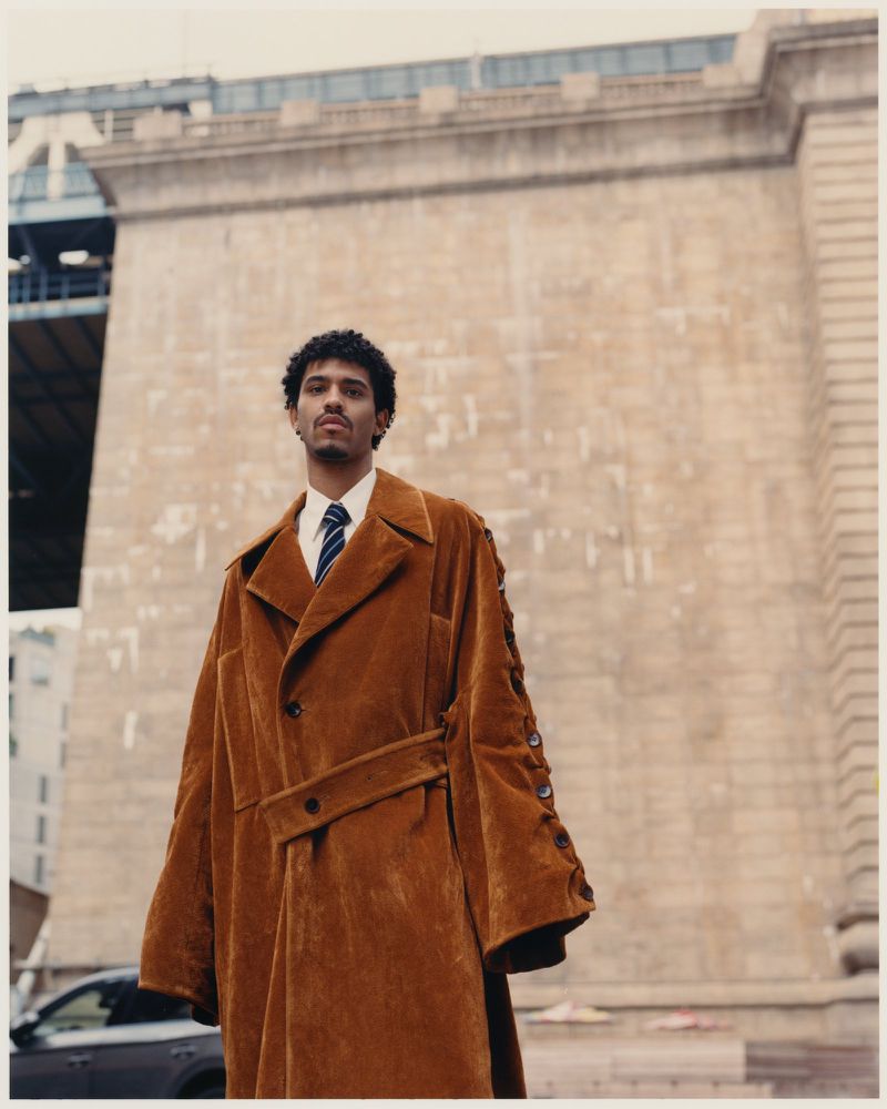 Man in oversized brown coat stands against an urban backdrop of buildings and a bridge, showcasing fashion in an outdoor setting.