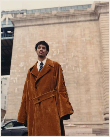 Man in oversized brown coat stands against an urban backdrop of buildings and a bridge, showcasing fashion in an outdoor setting.