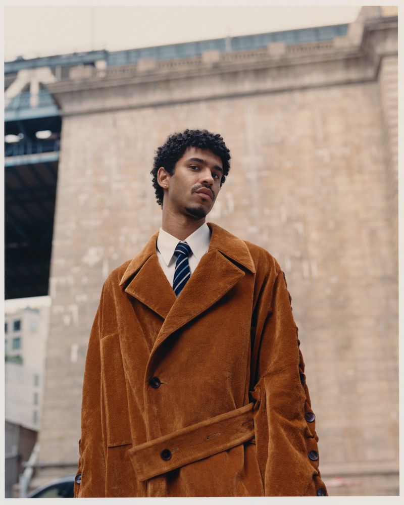 A young man wearing a brown corduroy coat and a striped tie outdoors against a stone bridge architectural background.