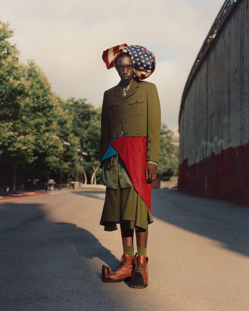 Fashion portrait of an African male model wearing a military-style green jacket, skirt, boots and an American flag-themed head covering outdoors.