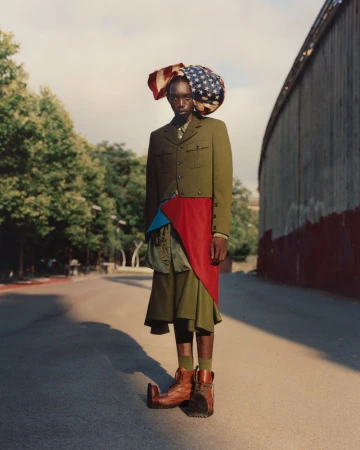 Fashion portrait of an African male model wearing a military-style green jacket, skirt, boots and an American flag-themed head covering outdoors.