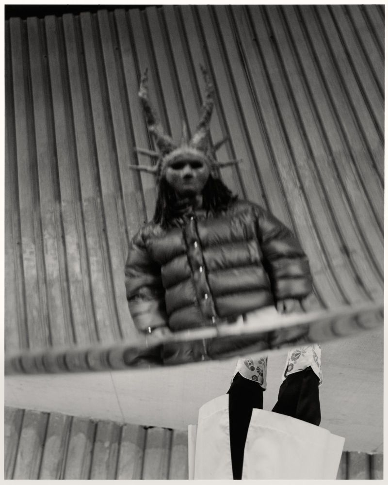 Person in a horned mask and padded jacket stands behind a partially visible railing, monochrome aesthetic and theatrical setting.