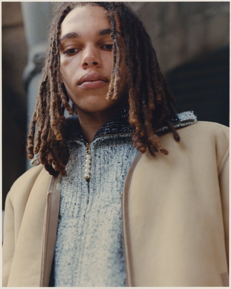 Young adult male poses in a three-quarter shot, wearing a sweater and jacket against a blurred urban background on an overcast day.