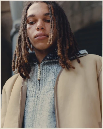 Young adult male poses in a three-quarter shot, wearing a sweater and jacket against a blurred urban background on an overcast day.