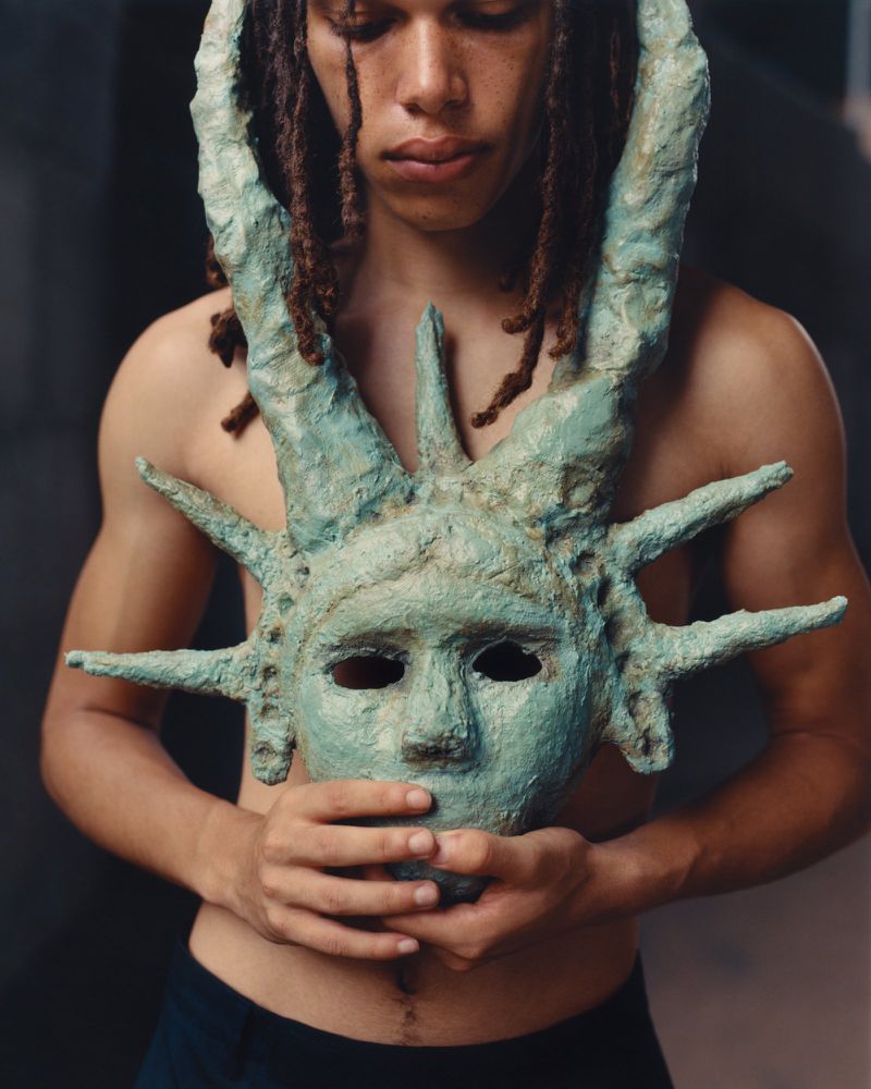 Person with dreadlocks holds a green statue mask, looking down in a close-up studio portrait with muted lighting and dark background.