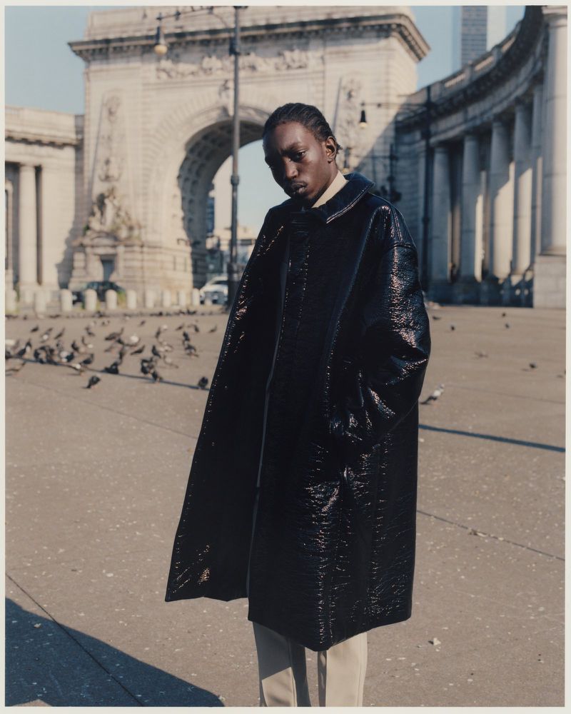 A young adult man in a glossy black coat stands in front of the Manhattan Bridge archway, with pigeons on the ground.