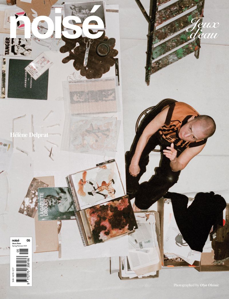 Overhead shot of a bald man posing near a white table covered with art books, paint supplies, and a metal ladder in an indoor setting.