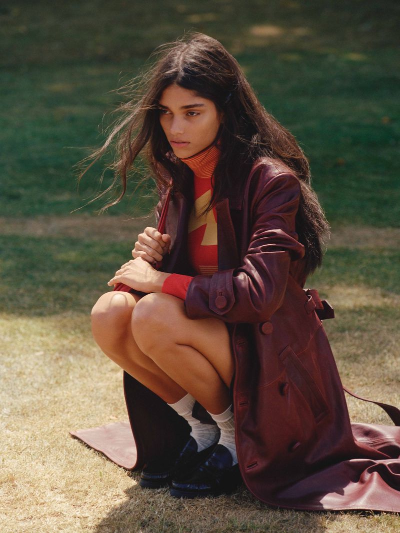 A brunette woman in a burgundy leather coat poses in a grassy field wearing a patterned top and white socks, sitting on a blanket.