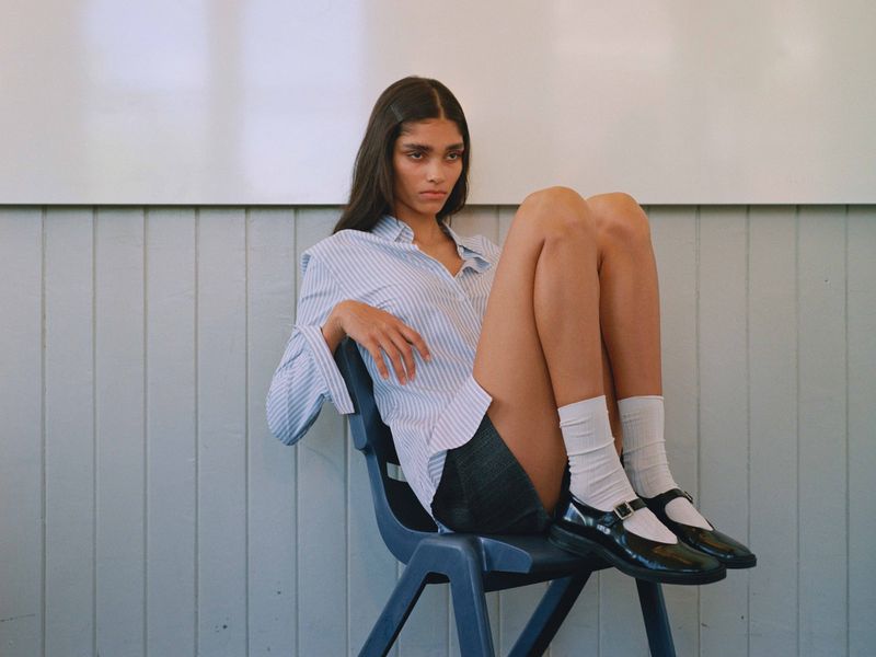 Woman sitting on blue plastic chair indoors. She is wearing a striped blouse, dark shorts, white socks, and black Mary Jane shoes.