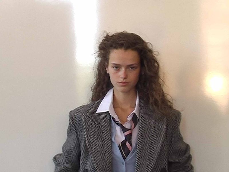A young woman with curly brown hair, wearing a school uniform-inspired outfit, stands in front of a plain white wall in a studio setting.