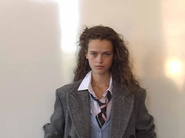 A young woman with curly brown hair, wearing a school uniform-inspired outfit, stands in front of a plain white wall in a studio setting.
