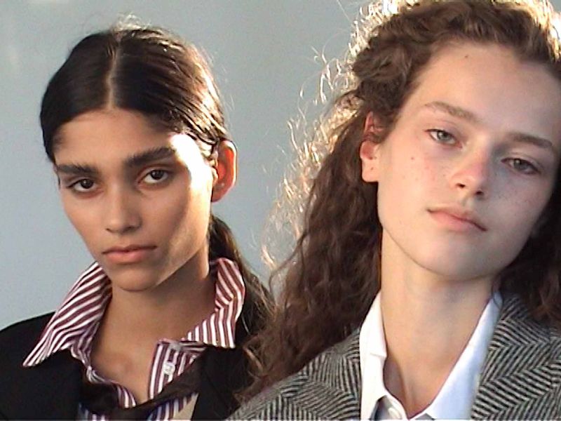 Two young female fashion models pose in a close-up studio shot, wearing jackets and button-down shirts, with one having dark hair and the other with light brown curly hair.