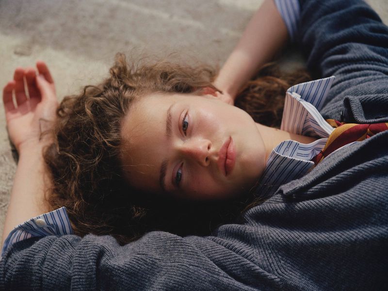 Young woman with curly hair lies on a tan surface, wearing a striped shirt, grey sweater, and tie in a close-up shot, low angle.