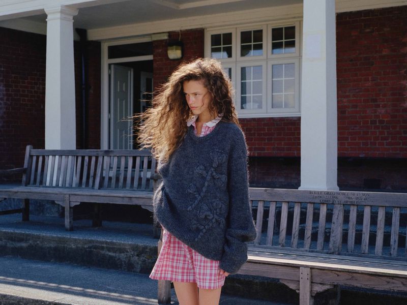 Young adult woman stands in front of a brick building wearing a gray sweater and red plaid skirt. Natural light.