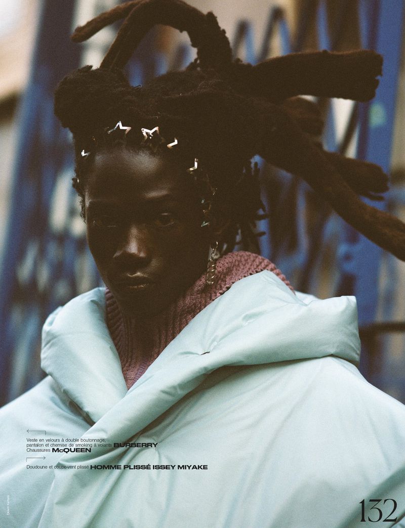 Fashion portrait of an adult model with dark skin and locs with accessories, wearing a light blue puffer coat in an urban environment.