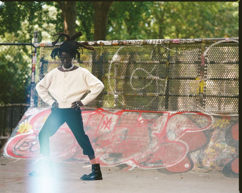 An adult with locs poses outdoors in a skatepark, wearing a cream sweater, dark pants, and black boots, against a backdrop of graffiti art.