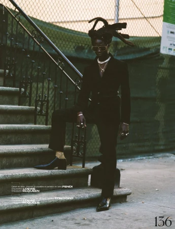 A stylish Black model in a dark suit and gold accessories poses on stairs outdoors, framed by a fence and neutral urban colors.