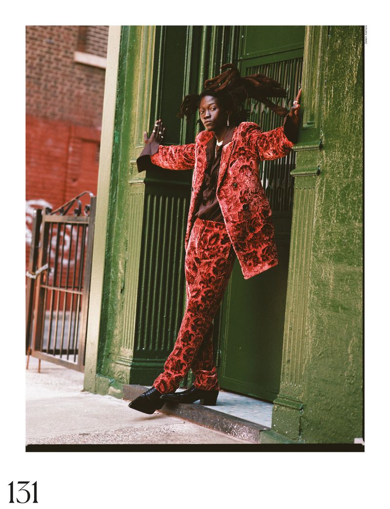 Woman in red floral suit posing by a green doorway, leaning with hands against the door frame, with a brick building in background.
