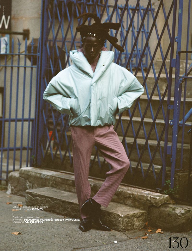 Fashion editorial featuring a Black model in a light blue puffer jacket and pink trousers standing against a blue gate on an urban street.