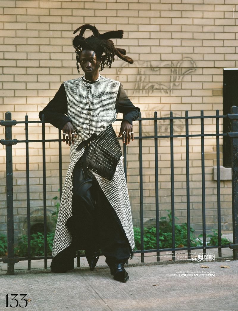 A black male model poses in a textured coat and black clothing against a brick wall, wearing rings, hair styled in dreadlocks, outdoors.