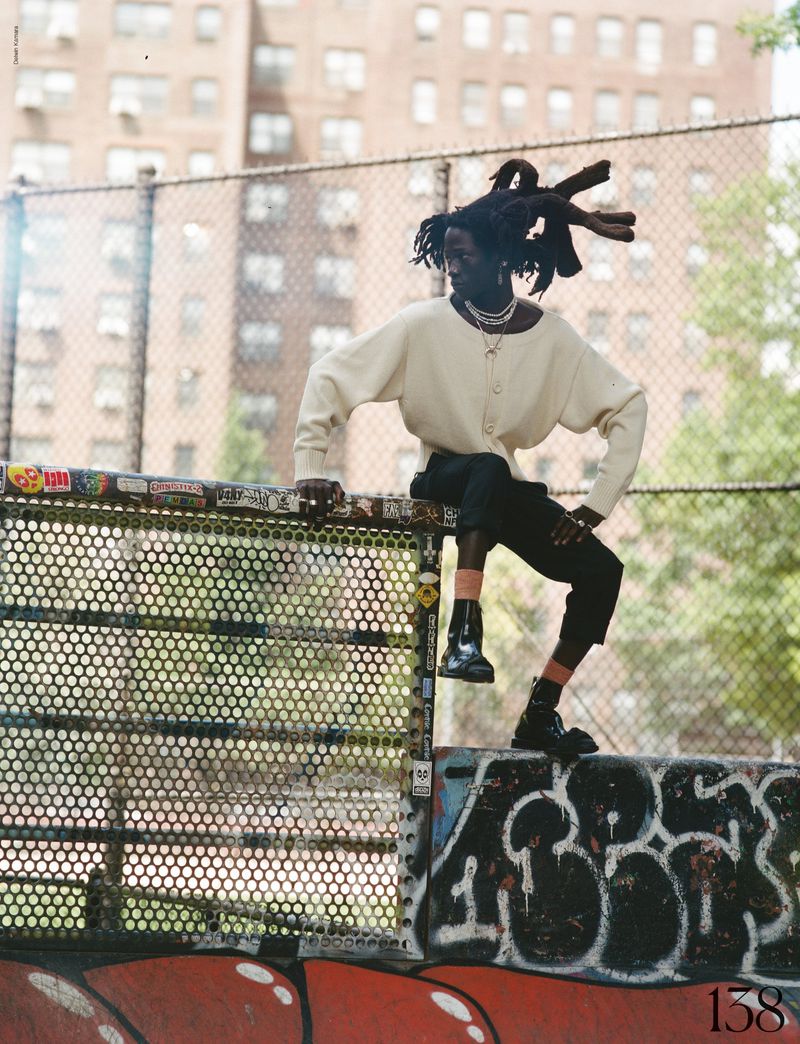 Adult male with dreadlocks poses on a park fence in an urban setting, wearing a cream-colored sweater and black pants with shiny black boots.