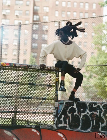 Adult male with dreadlocks poses on a park fence in an urban setting, wearing a cream-colored sweater and black pants with shiny black boots.