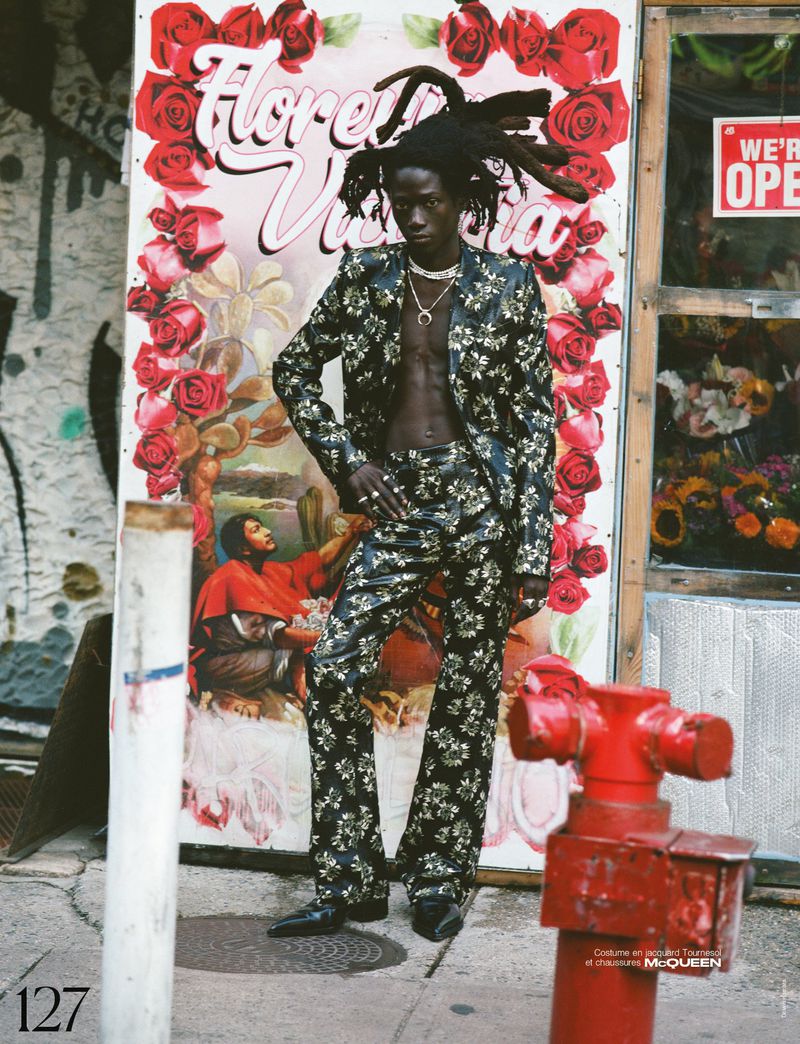 Dark-skinned male model in floral suit poses in front of flower shop advertisement and red fire hydrant on city street, editorial fashion.