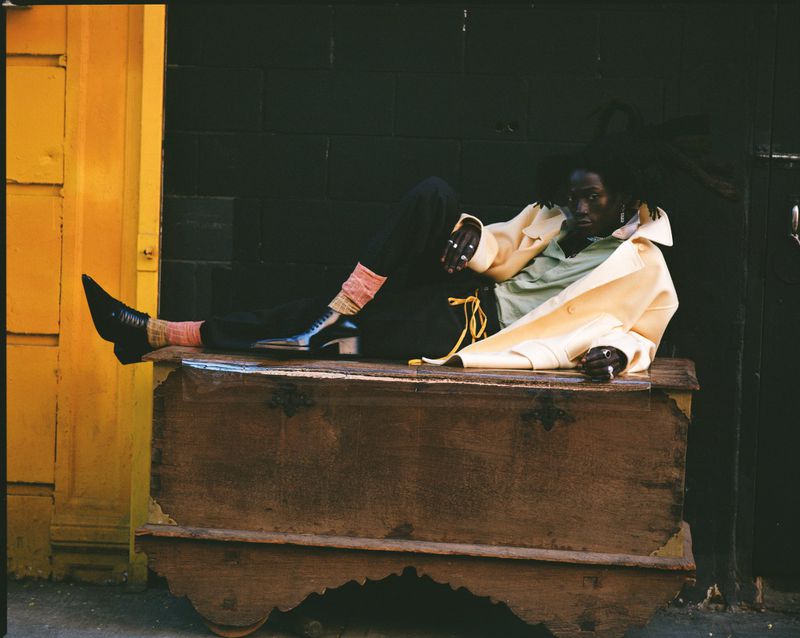 Person with dark skin and dreadlocks wearing a tan jacket and black pants, sitting on a wooden chest in front of a yellow door.