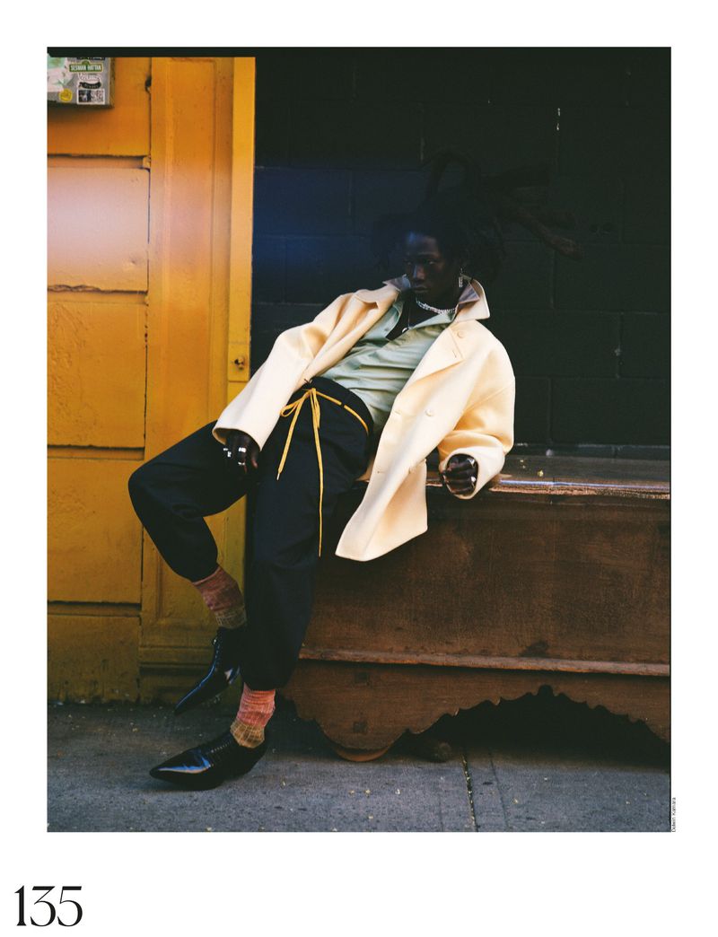 An adult Black male sits on a ledge in black pants, shiny shoes, and a cream jacket with a light green shirt. Urban setting.