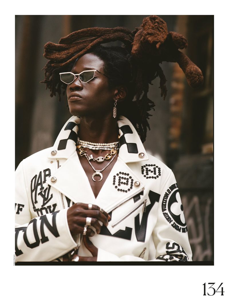 Portrait of a stylish adult male model wearing sunglasses and a white jacket with dreadlocks, posing against an urban background.