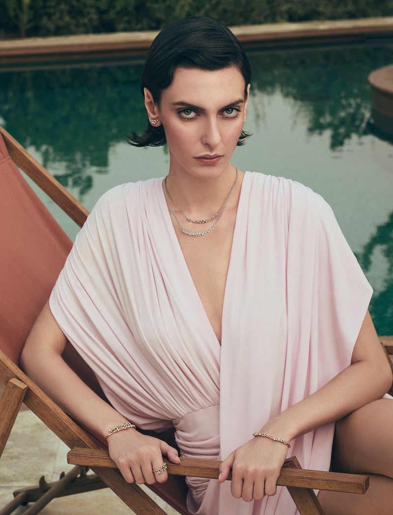 Elegant woman with short dark hair poses outdoors in a pink dress and jewelry, sitting by a swimming pool on a sunny day.