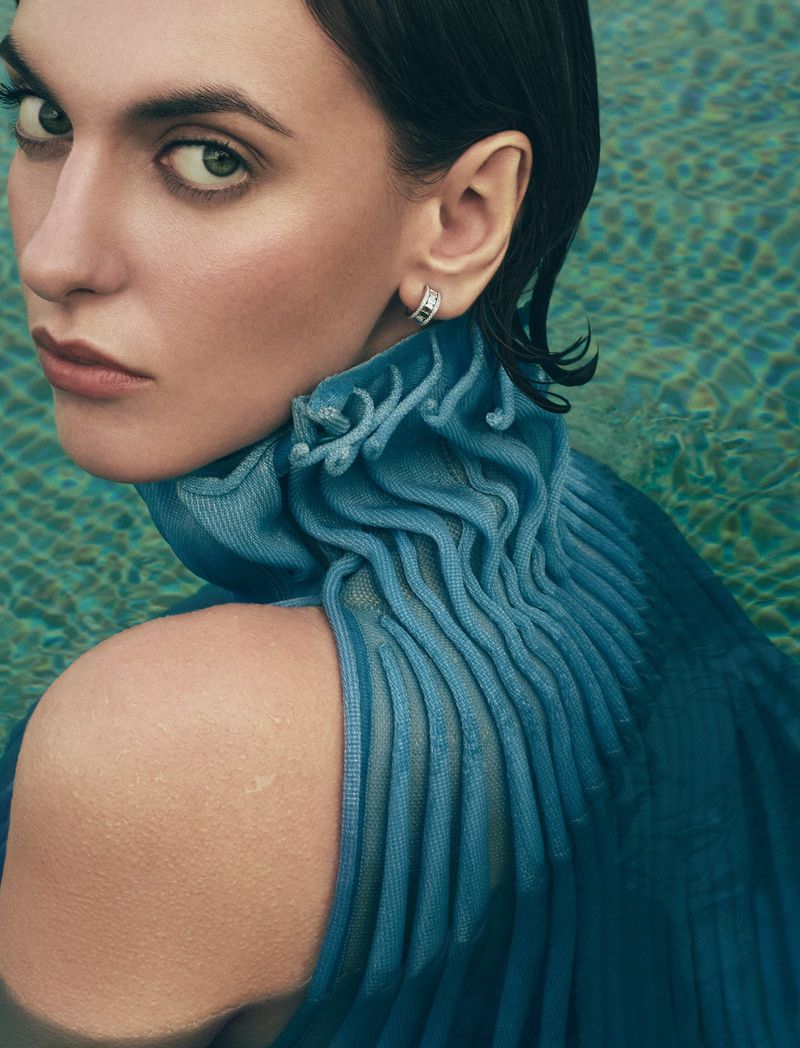 Woman with short dark hair, wearing a ruffled blue top and silver hoop earring, poses in front of shimmering water creating a fashion portrait.