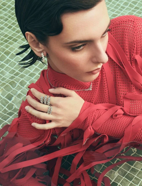 Woman in red mesh top with wet hair and jewelry, including rings, earrings, and a necklace, poses against a green tiled backdrop.
