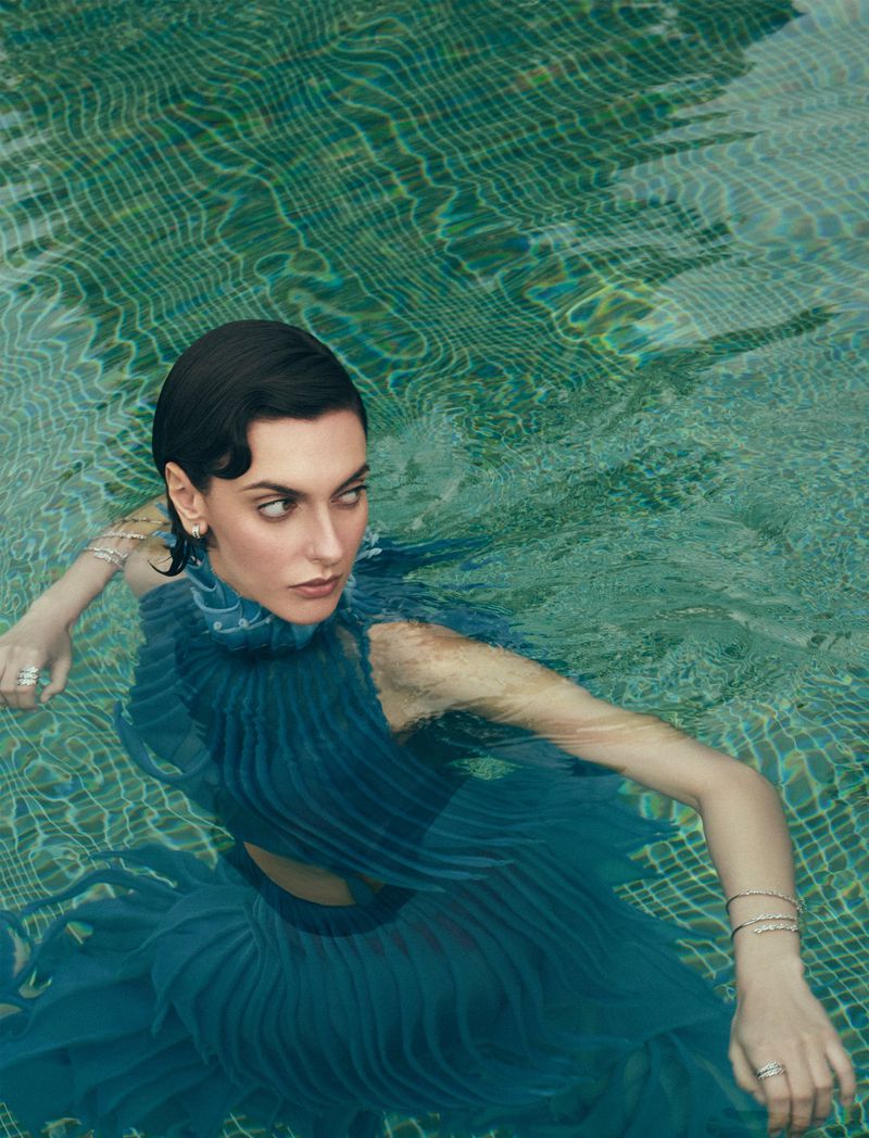 Woman with short dark hair wearing a blue dress, in water with tile details, rings, and bracelets, looking directly at the viewer.