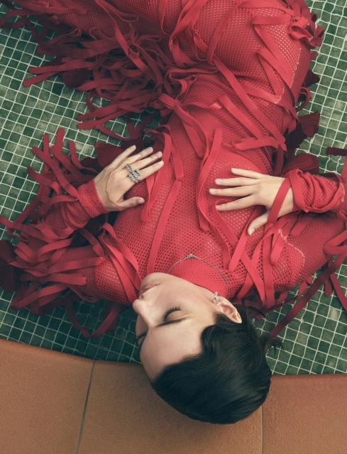 Woman in a red dress with fringe lying on a tile floor with green and white grid, head resting on a terracotta tile, jewelry visible.