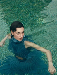 Woman in a teal pleated dress submerged in an outdoor pool wearing diamond bracelets