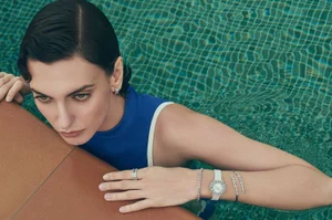 Woman in blue swimwear resting arms on a pool edge, wearing diamond jewellery and a watch