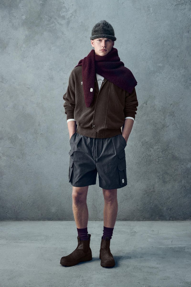 Full shot of a male model in a brown zip jacket, gray shorts, brown beanie and boots, and a burgundy scarf against a grey backdrop.