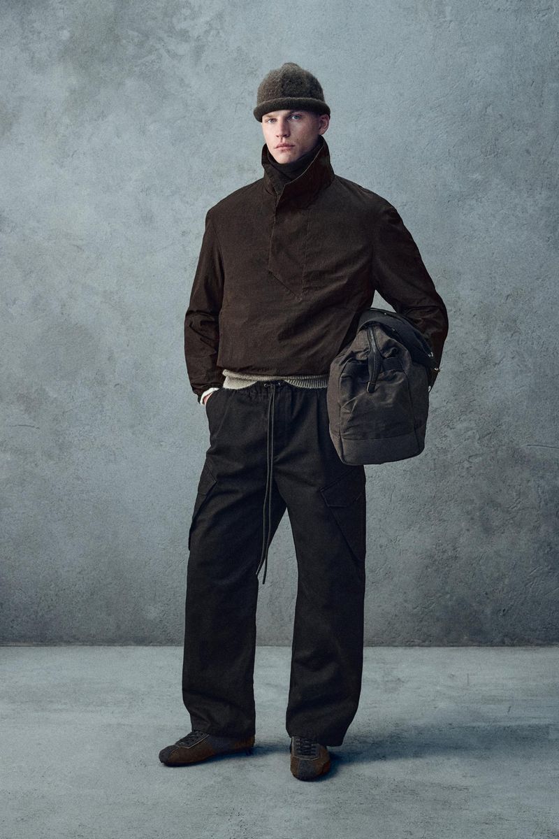 Male fashion model poses in a brown jacket, black pants, and beanie with a shoulder bag against a neutral grey studio backdrop.
