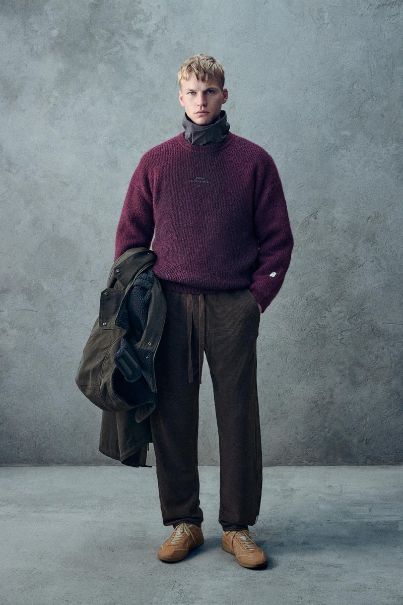 Man in a burgundy sweater and brown corduroy pants stands against a grey wall, holding a jacket, showcasing a fashion editorial style.