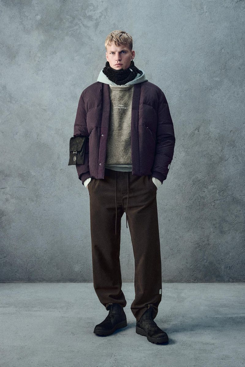 A full shot of a blonde male model posing in a studio, wearing a mauve puffer jacket, grey hooded sweatshirt, brown pants, and black boots.