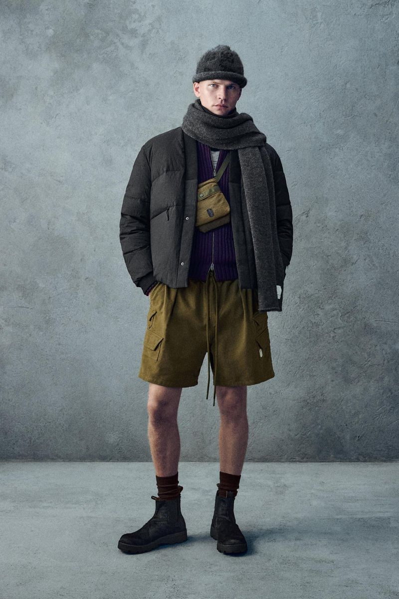 An adult male is standing indoors against a textured wall, wearing a beanie, scarf, puffer jacket, cargo shorts, and boots, looking forward.