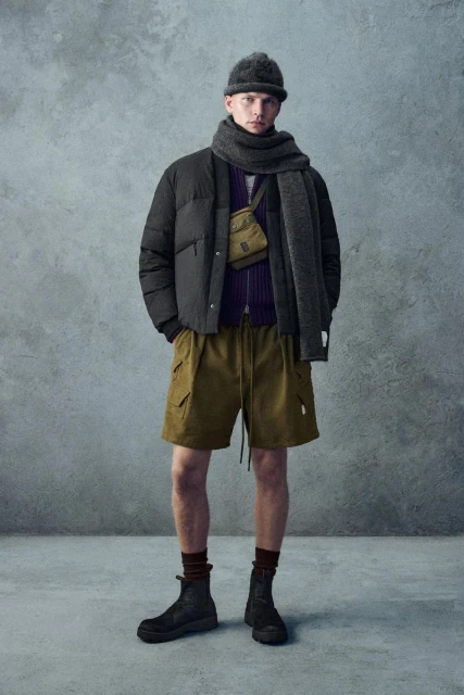 An adult male is standing indoors against a textured wall, wearing a beanie, scarf, puffer jacket, cargo shorts, and boots, looking forward.