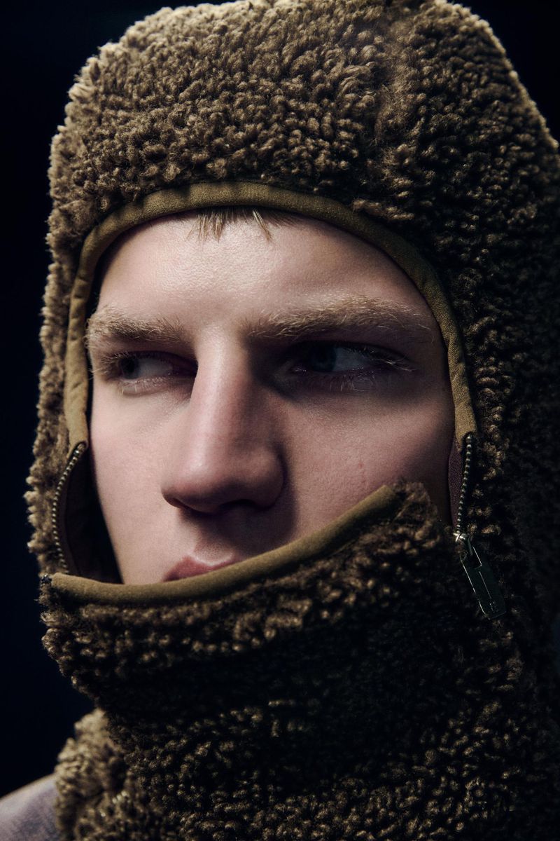 Close-up of a light-skinned adult male with blond hair wearing a fuzzy brown balaclava against a dark background in a studio setting.