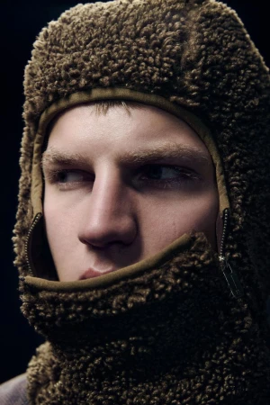Close-up of a light-skinned adult male with blond hair wearing a fuzzy brown balaclava against a dark background in a studio setting.