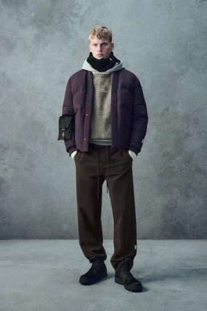 Full-length portrait of a male model wearing a quilted puffer jacket and layered hoodie against a textured grey studio wall background.